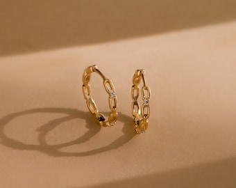 May include: A pair of gold-coloured hoop earrings. Each earring has a chain-link design with small, clear gemstones. The earrings are displayed on a neutral, light brown surface, with a soft shadow.