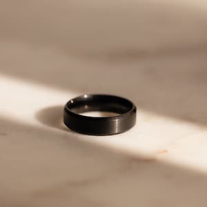 Puede incluir: Un anillo negro con acabado cepillado. El anillo es de metal y tiene un diseño sencillo.