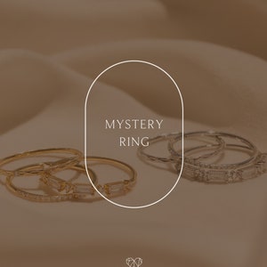 May include: A set of four gold and silver rings with clear stones. The rings are stacked on top of each other and the text "MYSTERY RING" is written in a white oval shape above the rings.