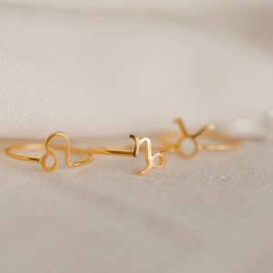 Könnte beinhalten: Vier goldene Ringe mit Sternzeichen. Die Ringe liegen auf einem weißen Stoffhintergrund. Die Zeichen sind Löwe, Steinbock, Stier und ein viertes Zeichen, das nicht sichtbar ist.