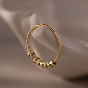 Może przedstawiać: Złoty pierścionek z pięcioma małymi, cylindrycznymi koralikami. Każdy koralik ma wygrawerowane imię czarną, stylizowaną czcionką. Pierścionek jest umieszczony na miękkim, neutralnym tle, podkreślającym design biżuterii.