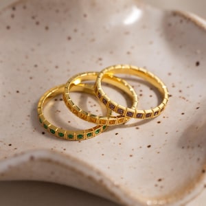 Puede incluir: Tres anillos dorados con piedras preciosas de colores se exhiben sobre un plato de cerámica moteado de color blanquecino. Cada anillo presenta una banda de pequeñas piedras cuadradas verdes, amarillas y moradas. Los anillos están apilados, mostrando su diseño.