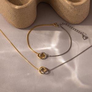 Puede incluir: Un conjunto de joyas en tonos dorados y plateados, que incluye un collar y una pulsera. Cada pieza presenta una cadena delicada y un colgante de doble círculo. La pulsera tiene una cadena ajustable con un dije de corazón.