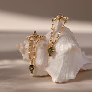 Puede incluir: Dos collares dorados con un diseño de estrella y un colgante de piedra verde. Los collares se exhiben sobre una concha blanca, con un fondo suave y neutro. Los colgantes tienen forma de diamante.