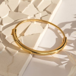 A close up of our Aris Hinge Bracelet in 18K gold placed on top of a white textured tile and white tabletop - featuring an about 73mm x 59mm and about 6mm thick statement bangle with a Hinge clasp design.