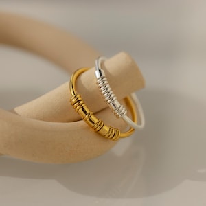 Könnte beinhalten: Zwei schlichte, minimalistische Ringe. Ein Ring ist golden, der andere silbern. Beide Ringe bestehen aus einer Reihe kleiner, runder Perlen.