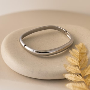 May include: A silver-coloured, curved bangle bracelet with a clasp closure. The bracelet is displayed on a light beige, oval-shaped stone, with a dried, golden-brown leaf in the lower right corner. The bracelet has a smooth, polished finish.