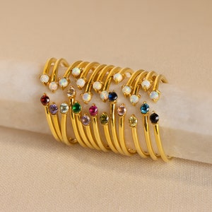 Könnte beinhalten: Ein Stapel goldener Ringe mit verschiedenen farbigen Edelsteinen, darunter Opal, Amethyst, Smaragd, Rubin, Saphir und Türkis.