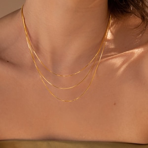 May include: A layered gold chain necklace with a delicate box chain design. The necklace is worn by a person with a light complexion.