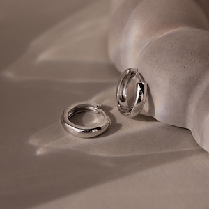 May include: Two silver-colored hoop earrings. The earrings are round and have a smooth, polished surface. The earrings are displayed on a neutral-toned surface, with soft lighting creating highlights and shadows. The overall aesthetic is minimalist and elegant.