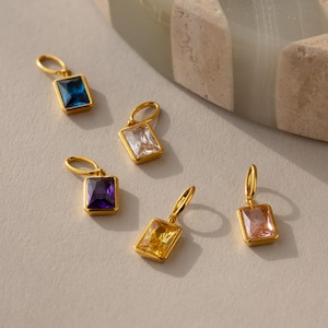 Op de afbeelding: Vijf gouden bedels met verschillende vierkante edelstenen in verschillende kleuren. De bedels liggen op een wit oppervlak.