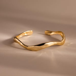 以下が含まれることがあります： 有機的で流れるようなデザインのゴールドカラーのカフブレスレット。 オープンエンドのブレスレットは、滑らかで研磨された仕上げと、光を反射するわずかに不規則な形状をしています。 ジュエリーはニュートラルな背景に表示され、その金属的な輝きを強調しています。