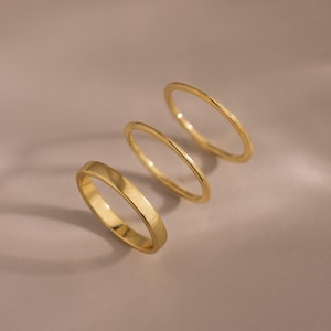 May include: Three gold rings of varying designs are displayed against a neutral background. The rings are thin and minimalist, with one featuring a flat, rectangular band, and the others are simple, rounded bands. The rings are made of a shiny, yellow gold.