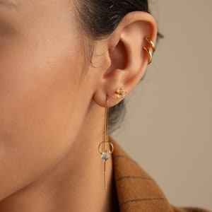 May include: Gold chain earrings with a clear crystal at the end of the chain. The earrings are hanging from the earlobe and have a small clear crystal at the top of the chain.