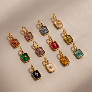 May include: A collection of gold-colored rectangular charms with a starburst design. Each charm features a different colored gemstone, including red, blue, green, purple, and white. The charms are displayed on a neutral background.