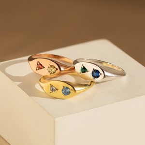 Könnte beinhalten: Drei Siegelringe mit verschiedenen Edelsteinen. Die Ringe sind übereinander gestapelt. Der oberste Ring ist silber mit einem blauen Edelstein, der mittlere Ring ist gold mit einem blauen und weißen Edelstein und der unterste Ring ist roségold mit einem roten und gelben Edelstein.