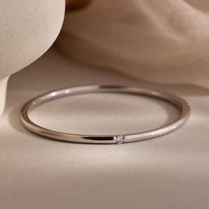 Könnte beinhalten: Ein silberfarbenes Armreif-Armband mit einem kleinen, klaren Edelstein, der in das Band eingesetzt ist. Das Armband ist rund und glatt und reflektiert das Licht. Der Schmuck wird auf einem neutralen Hintergrund präsentiert.