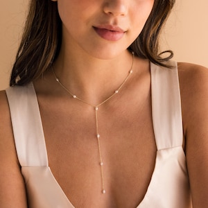 Puede incluir: Un collar de cadena dorada con pequeñas perlas blancas. El collar es de estilo lariat, con una caída larga que cuelga en la parte delantera del cuello.