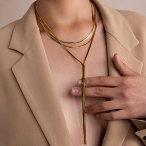 May include: A gold layered necklace with a long, thin pendant. The necklace is worn by a person with a tan blazer.