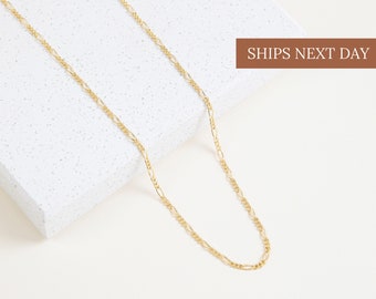 May include: A gold-colored Figaro chain necklace. The necklace is displayed on a white textured surface. The text "SHIPS NEXT DAY" is visible in a brown rectangle in the upper right corner.