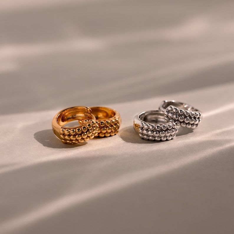 May include: Two pairs of hoop earrings, one gold and one silver, are displayed. The earrings have a textured surface of small, evenly spaced spheres. The gold earrings are positioned in front of the silver earrings.