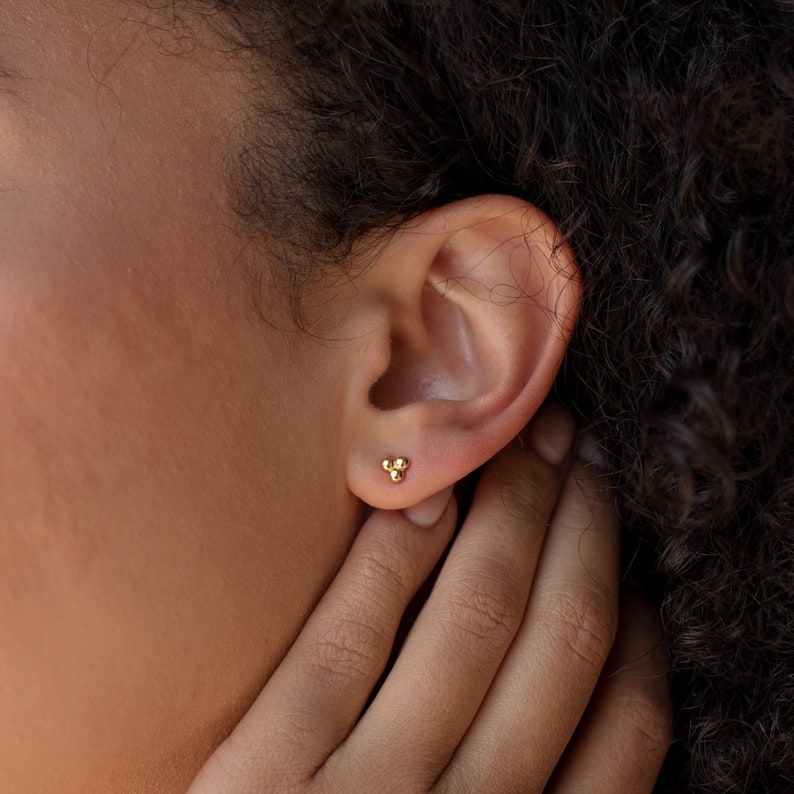 May include: A close-up of a person's ear with a small gold stud earring featuring three tiny balls.