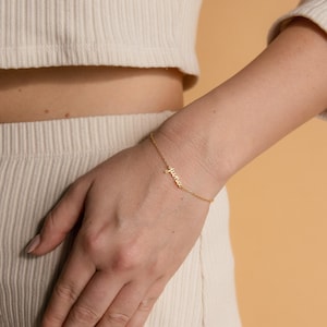 May include: A fine gold bracelet, inscribed with the word "june" in cursive script, is shown on a wrist. The bracelet's design is simple and elegant, with "june" as the focal point. The background has a soft, warm tone.