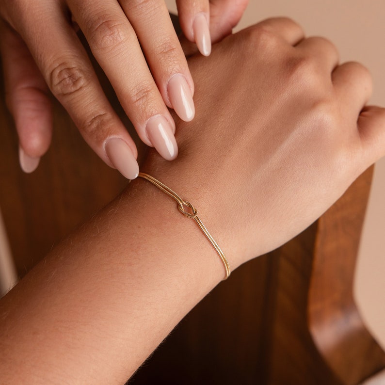 May include: A gold bracelet with a knot design. The bracelet is on a person's wrist.