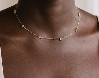 Op de afbeelding: Een goudkleurige ketting met een fijne schakelketting versierd met gelijkmatig verdeelde, kleine bloemhangers met zes bloemblaadjes. De metalen ketting rust op de hals en toont het herhalende bloemmotief in de schakelstructuur.
