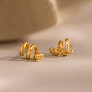 May include: A pair of gold-toned stud earrings shaped like coiled snakes. Each earring features a textured surface that mimics the appearance of snake scales, with the head of the snake forming the end of the spiral design in a polished finish.