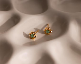 May include: A pair of gold stud earrings featuring a twisted metal design that forms a circular frame around a small, round, light green stone set in the centre of each piece, showcasing a polished metallic finish and a delicate, symmetrical structure.
