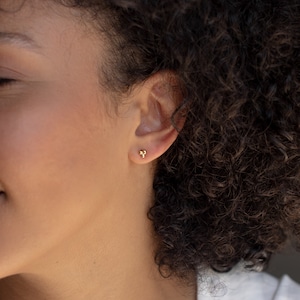 May include: A close-up of a person's ear with a small gold stud earring. The earring has three small, round beads.