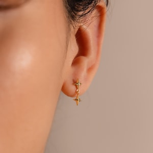 May include: A gold-toned earring featuring two four-pointed star shapes connected by a small link. The top star is fixed to the earlobe, while the bottom star hangs freely, creating a vertical, dangling design with a polished, reflective finish.
