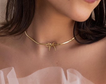 May include: A gold-toned choker necklace featuring a flat, flexible snake chain design. The center of the necklace is adorned with a small, polished gold-toned bow charm that sits horizontally against the base of the neck.
