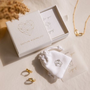 Puede incluir: Una caja blanca con un dise&ntilde;o de coraz&oacute;n geom&eacute;trico dorado y el texto "Caitlynn Minimalist" en la parte delantera. La caja est&aacute; abierta y contiene una peque&ntilde;a bolsa blanca con un dise&ntilde;o de coraz&oacute;n dorado y el texto "for you" y "enjoy" en una tarjeta blanca. La caja est&aacute; sobre una superficie blanca.