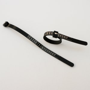 May include: Black plastic ring sizer with a flexible tape measure marked with numbers 1 to 17. The tape measure is attached to a ring sizer that can be adjusted to fit different finger sizes.