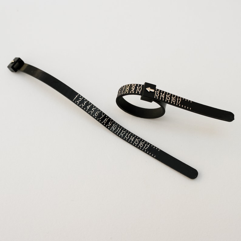 May include: Black plastic ring sizer with a flexible tape measure marked with numbers 1 to 17. The tape measure is attached to a ring sizer that can be adjusted to fit different finger sizes.