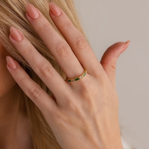 May include: A gold ring with a series of rectangular green and pink gemstones. The ring is worn on a finger, with the hand and part of the arm visible. The ring is a piece of jewelry.