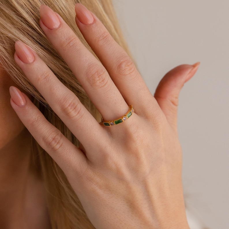May include: A gold ring with a series of rectangular green and pink gemstones. The ring is worn on a finger, with the hand and part of the arm visible. The ring is a piece of jewelry.