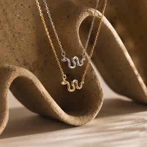 May include: Two delicate necklaces featuring small, serpentine-shaped pendants. One necklace is crafted in a silver-toned metal, while the other is in a gold-toned metal. Both snake pendants are embellished with tiny, clear, sparkling stones.
