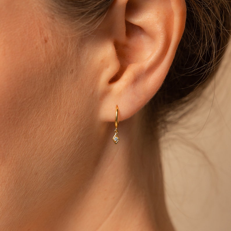 Close up on ear. A gold huggie hoop earring is in the the first lobe piercing. The hoop earring has a dangling charm in a dainty diamond shape with a circular diamond stone nestled inside it.