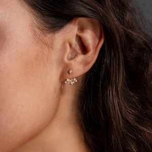 Può includere: Un orecchino ear jacket color oro caratterizzato da un piccolo perno a forma di stella sul davanti e da un gruppo curvo di ciondoli a forma di stella con pietre chiare incastonate che si posiziona sotto il lobo, creando un design a strati.