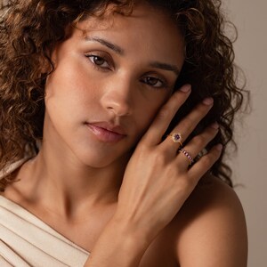 May include: A gold ring featuring a central purple stone surrounded by a halo of small clear stones, worn alongside a second gold ring set with a row of small pink stones. Both rings are displayed on a finger, showcasing their metallic bands.