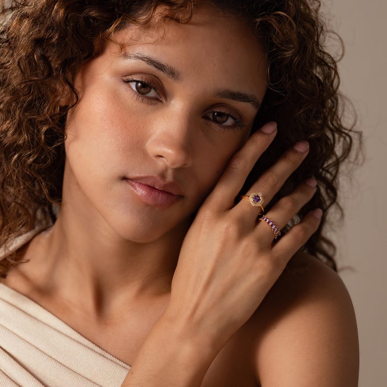 May include: A gold ring featuring a central purple stone surrounded by a halo of small clear stones, worn alongside a second gold ring set with a row of small pink stones. Both rings are displayed on a finger, showcasing their metallic bands.