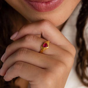 Puede incluir: Un anillo de tono dorado con una piedra preciosa roja en forma de corazón en el centro. La banda presenta un diseño acanalado y texturizado que se estrecha hacia el engaste, el cual sostiene firmemente la piedra roja vibrante y pulida.