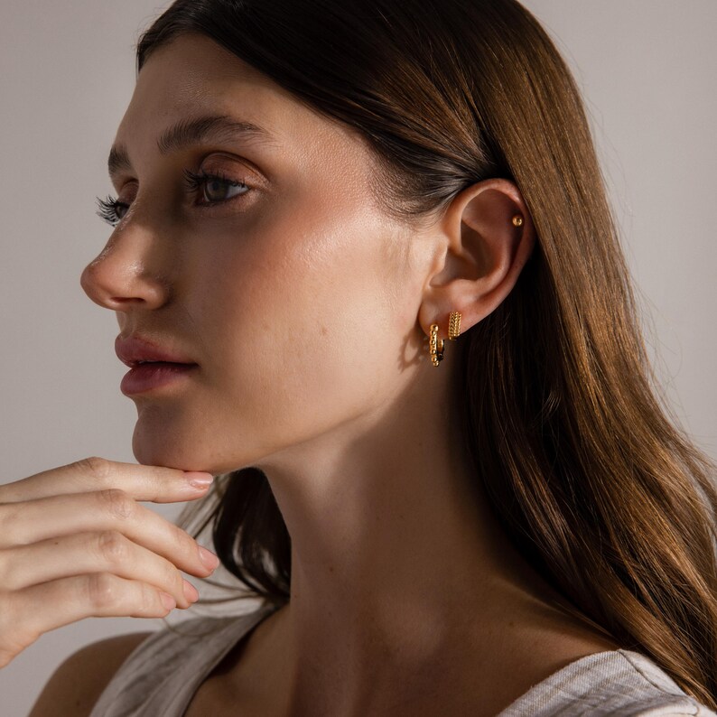 May include: A side profile of a person wearing two gold hoop earrings. The earrings are small and have a textured design. The person has long brown hair and is wearing a light-colored top.
