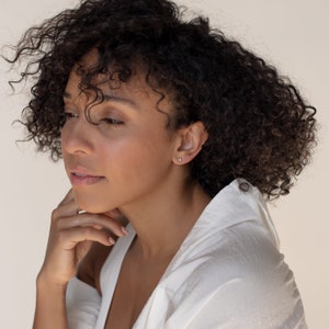 May include: A woman with curly brown hair wearing a white shirt. She is looking down and to the side. She is wearing a small stud earring in her left ear.