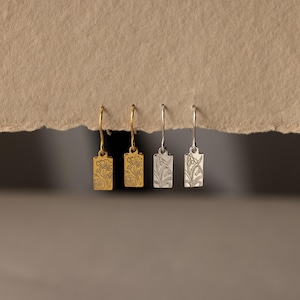 May include: Four rectangular drop earrings hanging in a row. The two on the left are gold-toned with an engraved floral design, while the two on the right are silver-toned and feature an engraved botanical pattern. Each earring has a thin hook.