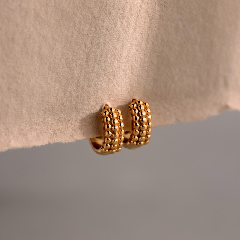 May include: A pair of gold-colored hoop earrings. The earrings have a textured surface with a series of small, round, raised bumps. The earrings are displayed against a neutral-toned background.
