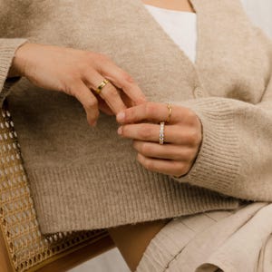 May include: Close-up of hands adorned with gold and diamond rings. The rings include a thick gold band, a delicate gold band, and a gold band with a row of small diamonds. The person is wearing a beige cardigan.